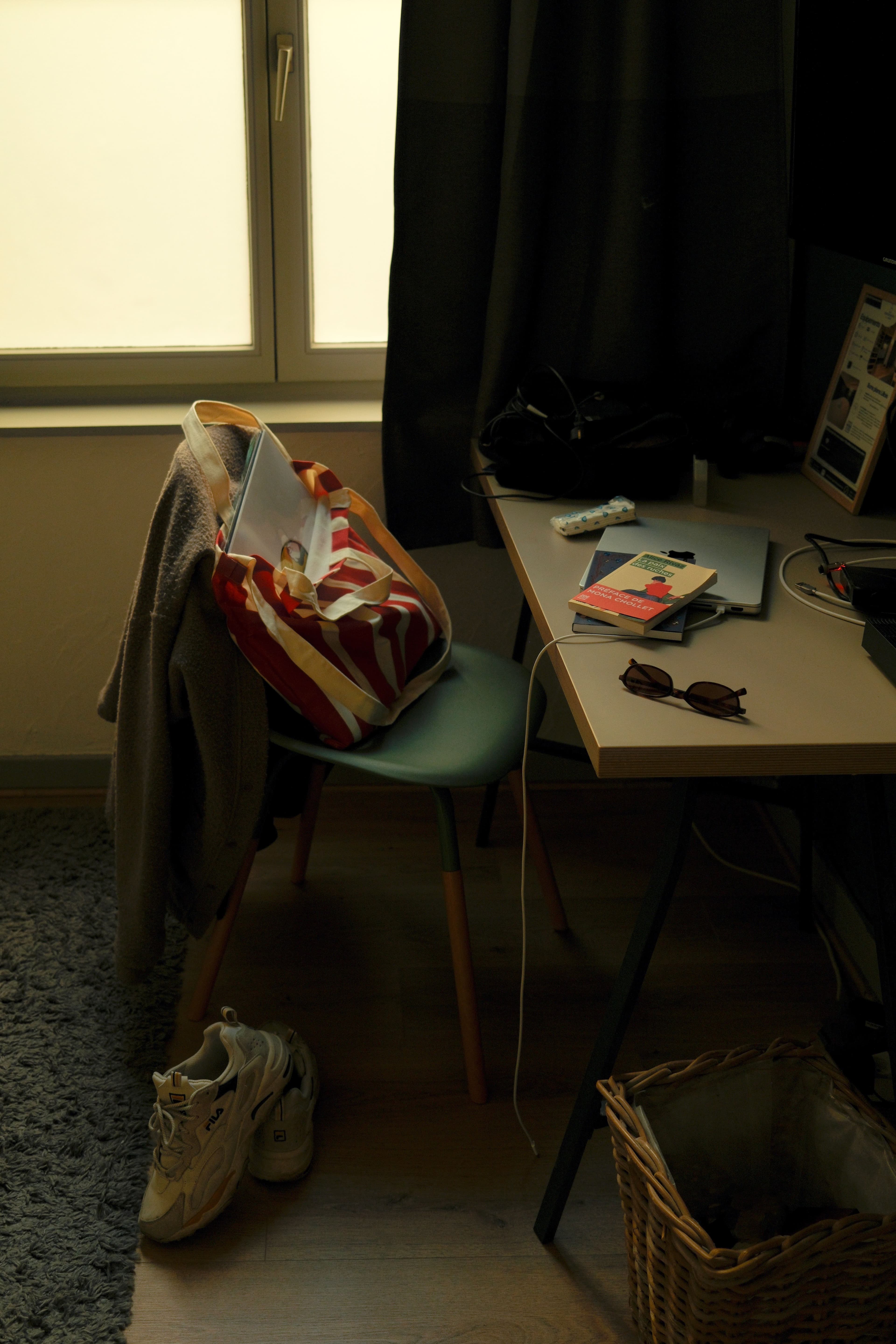 Desk and chair by a window, with a bag, coat, and sneakers nearby