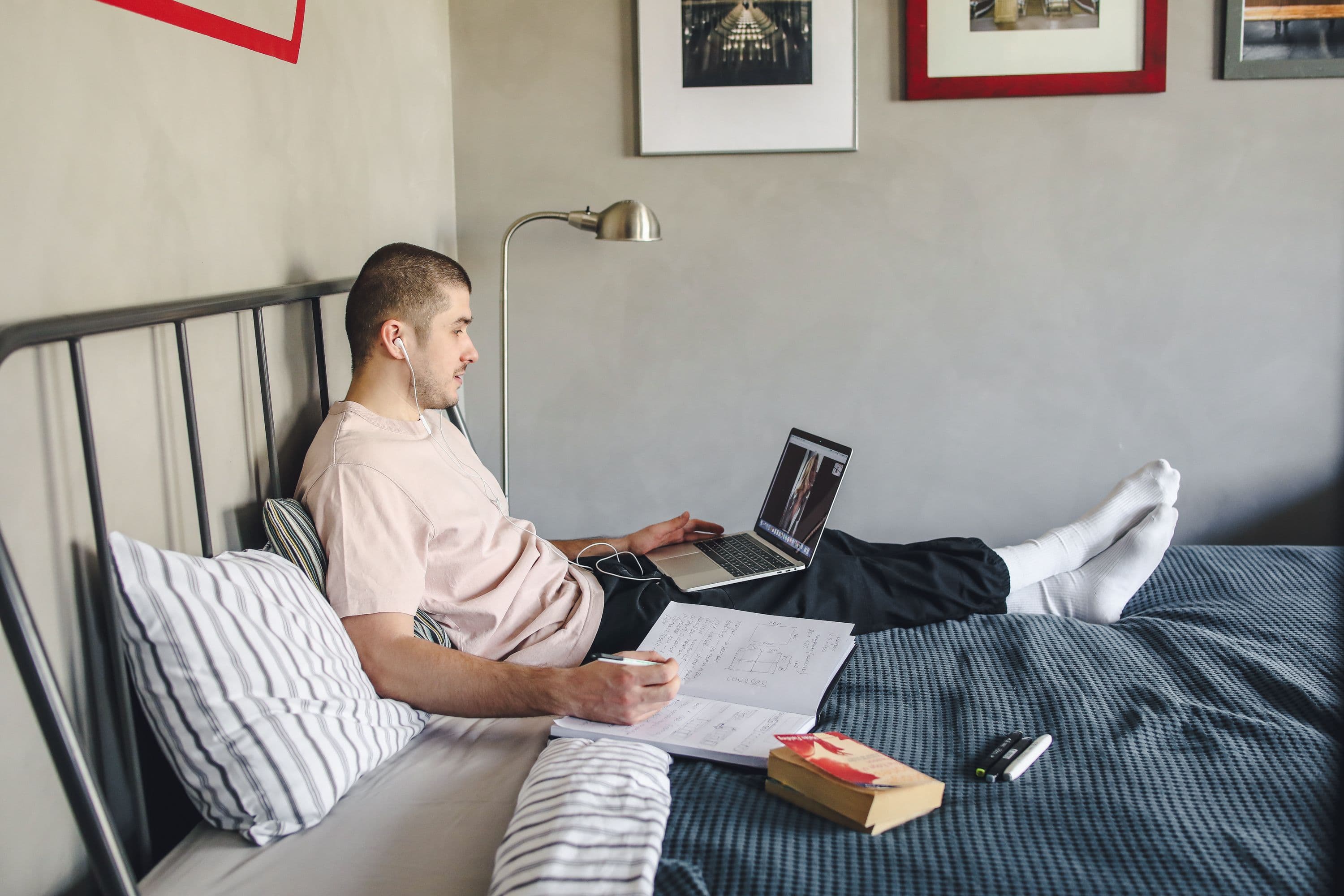 Student studying on a bed with a laptop, notebook, and floor lamp